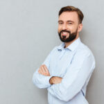 Pleased bearded man in business clothes with crossed arms looking at the camera over gray background