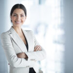 Closeup portrait of smiling beautiful middle-aged business woman wearing jacket and standing in light office hall with her arms crossed